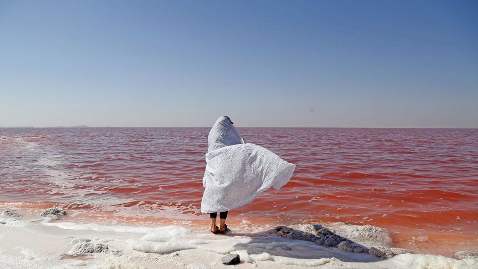 Person in a white shawl stands of the shoreline of Lake Urmia and its red-coloured water