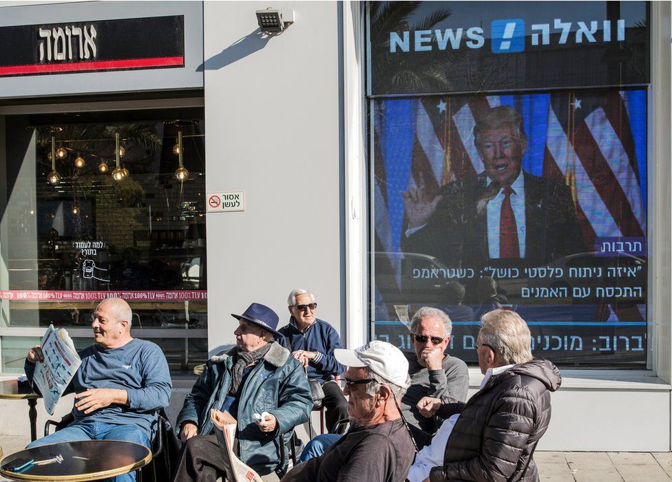 Israelis sit outside cafe in front of screen showing Donald Trump