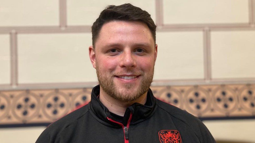 Man with brown hair and short beard wearing a black track suit