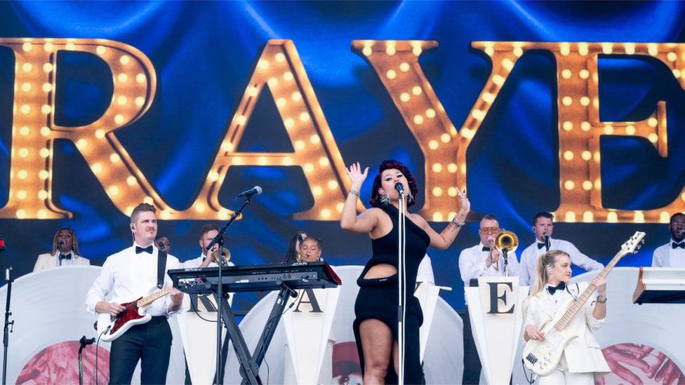 Raye plays Glastonbury. Her name is emblazoned in Gold behind her with an orchestra dressed in white in front of the sign. Raye, a woman, is wearing a black dress singing into a black microphone with her hands raised.