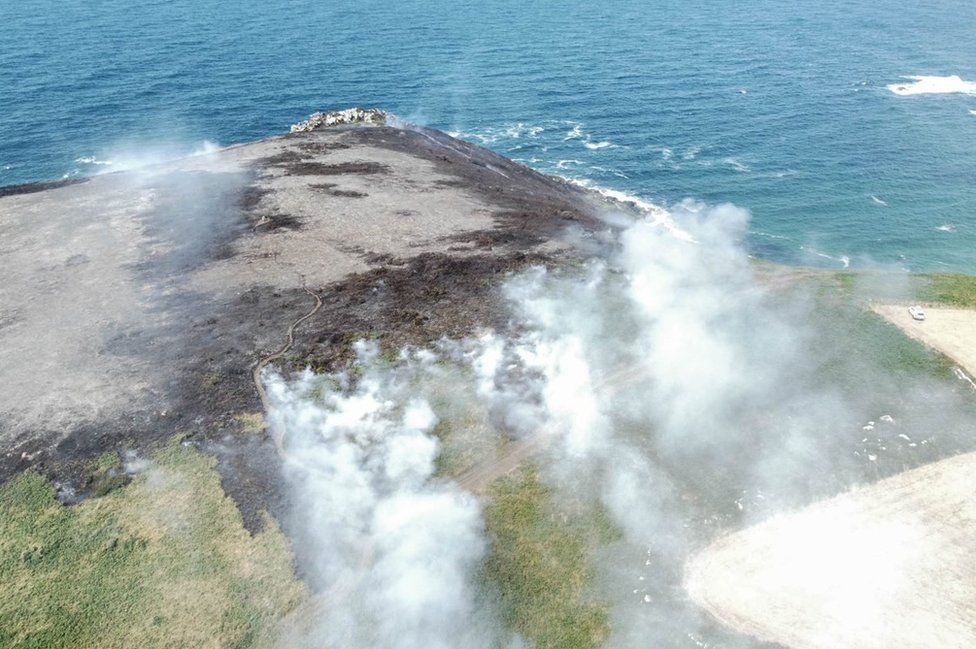 Aerial view of smoke and burnt patch of grass on cliff top next to sea