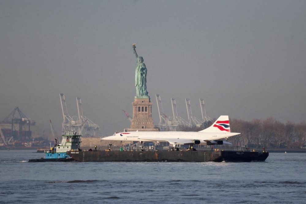 The Concorde supersonic jet is carried on a barge along the Hudson River past the Statue of Liberty