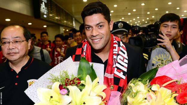 Hulk smiles as he walks through a crowded airport, carrying a bouquet of flowers while wearing a Shanghai SIPG scarf