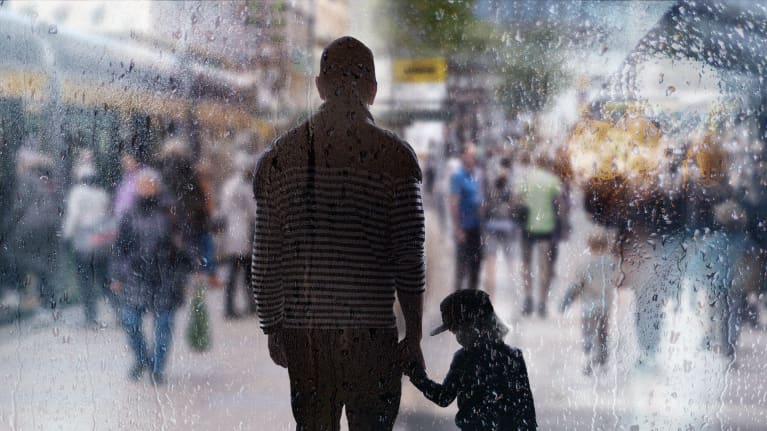 Mies seisoo lapsen kanssa käsikädessä vilkkaalla kadulla. Kuva otettu vesisateisen ikkunan läpi.