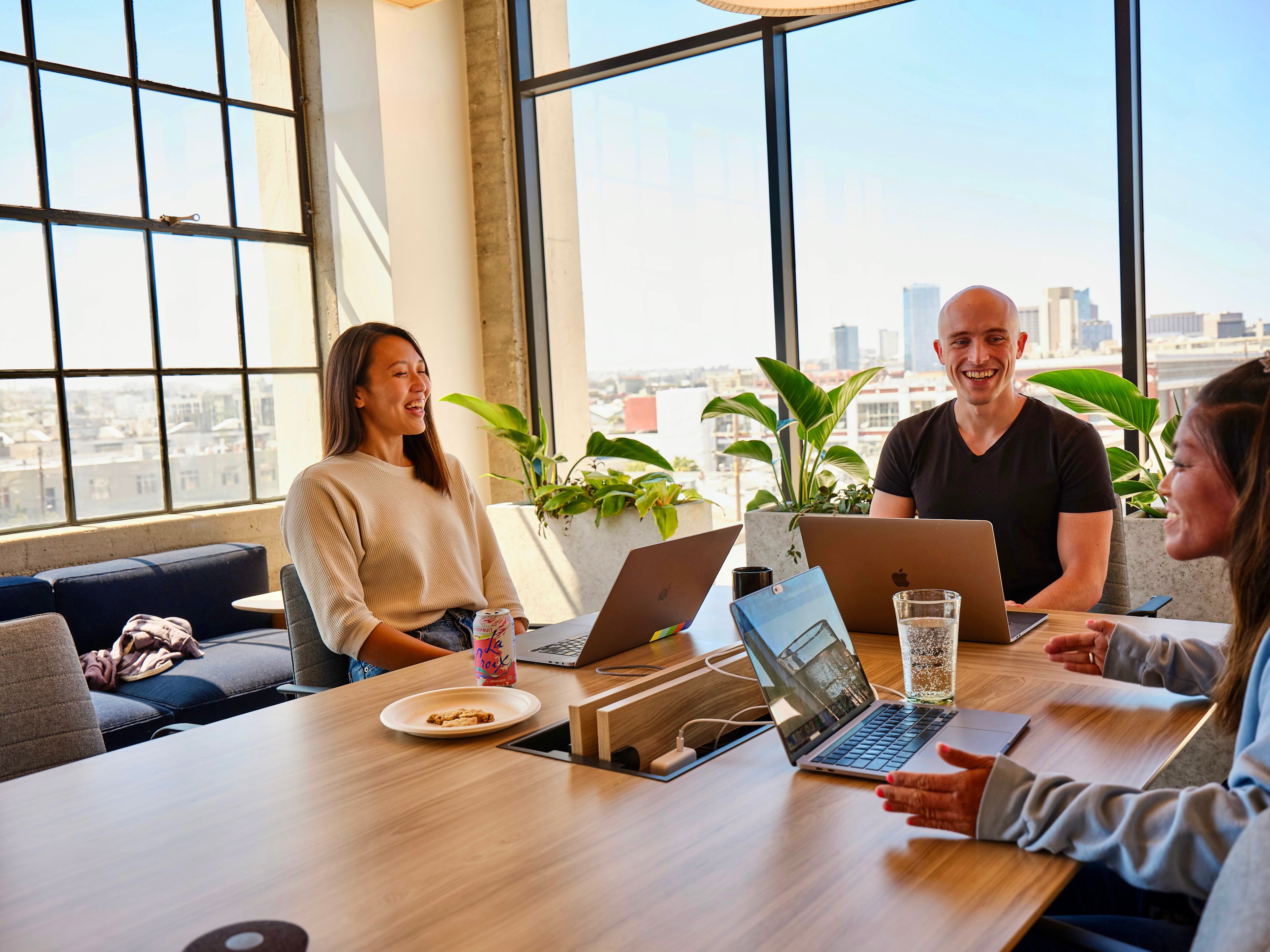 Three people sitting in a bright meeting room with large windows, smiling and talking around a large table with laptops in front of them