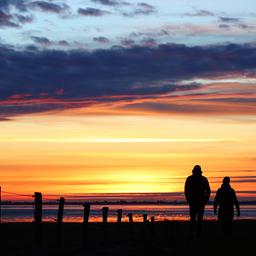 Zwei Menschen gehen vor einem farbenreichen Himmel kurz nach Sonnenuntergang an der Nordsee bei Husum spazieren.