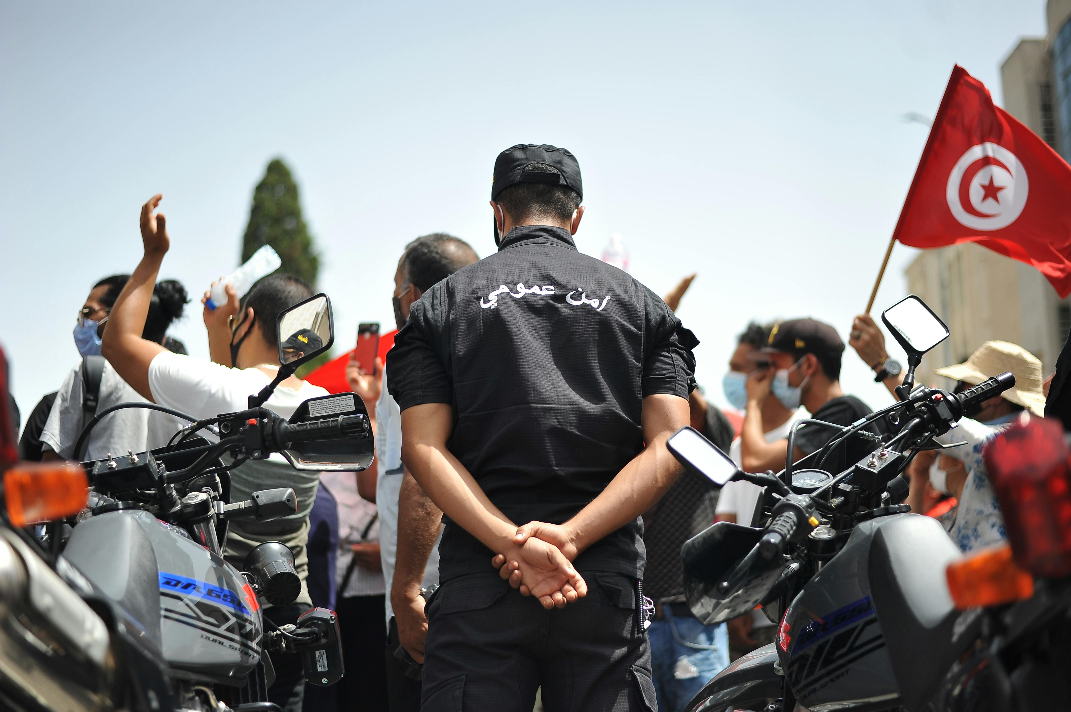 A police agent, framed by two motorcycles, turns his back to the picture as protestors carrying Tunisian flags walk by him.