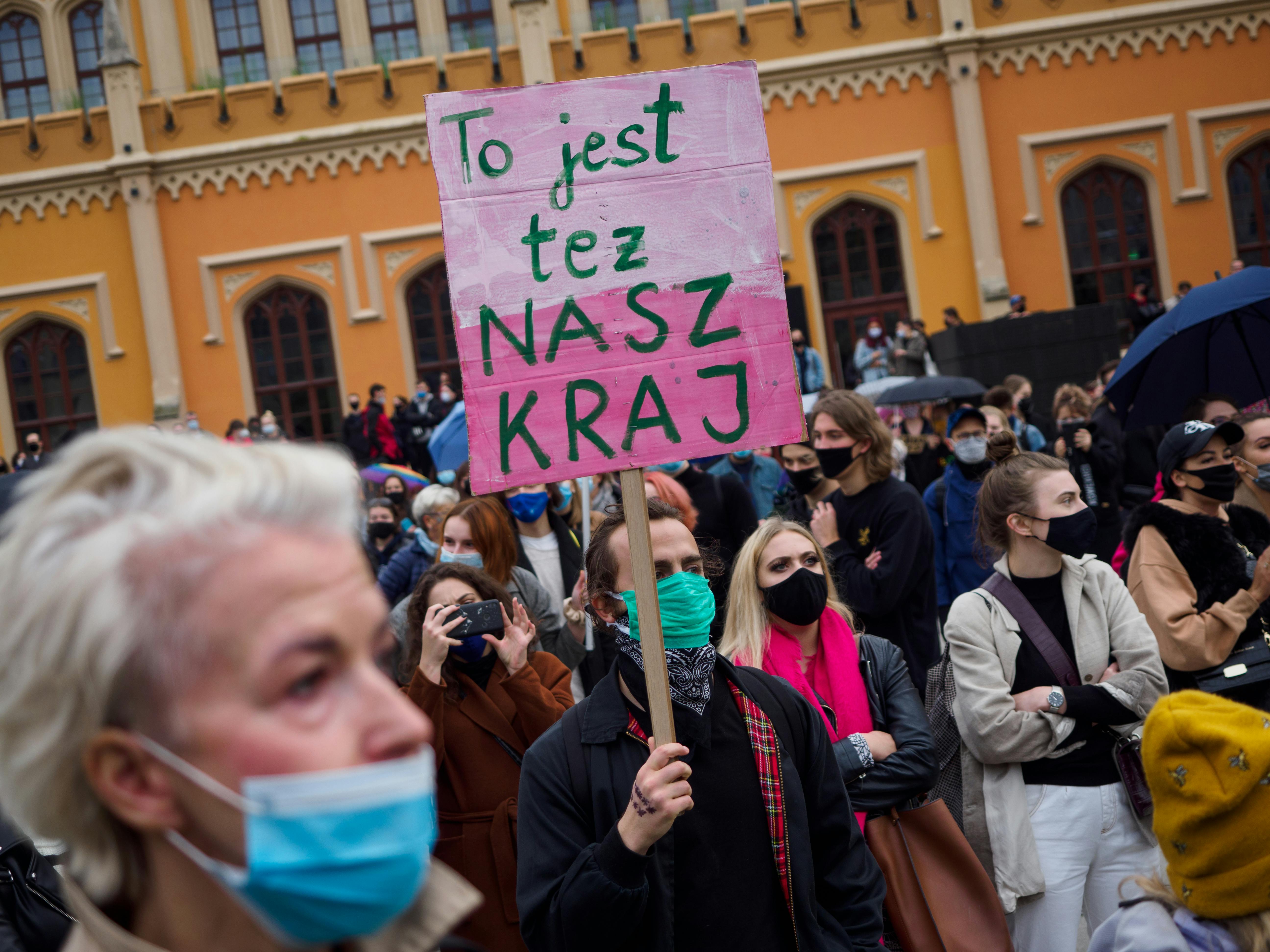 Protestors wearing masks gather in the streets of Poland. One holds a pink sign.