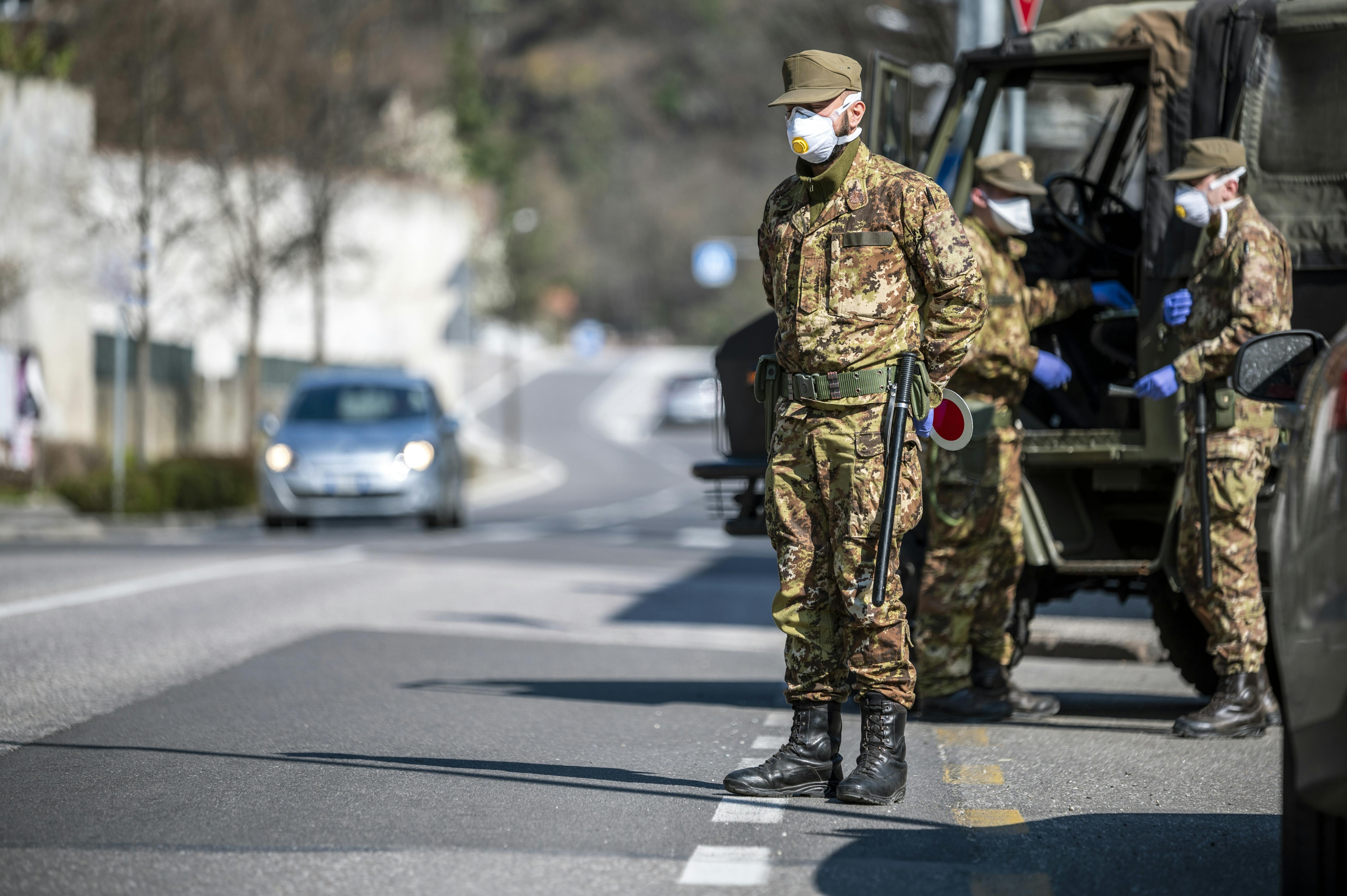 A man wearing military clothes an a mask stands before an army truck, on the side of the road. Behind him, two other soldiers are talking near the truck, wearing blue gloves and masks.