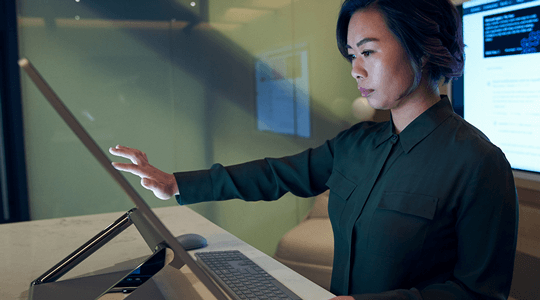 Side profile of a woman wearing a dark shirt in a dim office scrolling or working on a Microsoft Surface Studio.