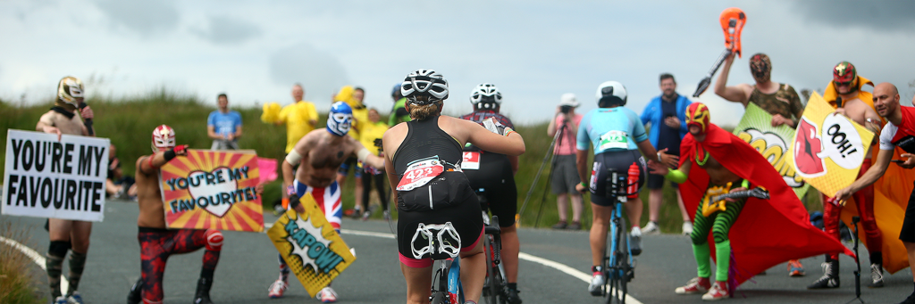 Athletes who are biking up a hill are motivated and supported by masked people carrying posters at IRONMAN UK