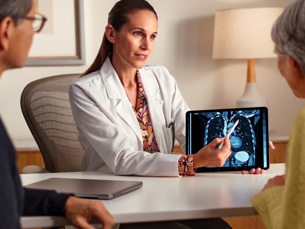 A female doctor in a lab coat pointing to an xray of lungs on a tablet while explaining something to two seated patients. 