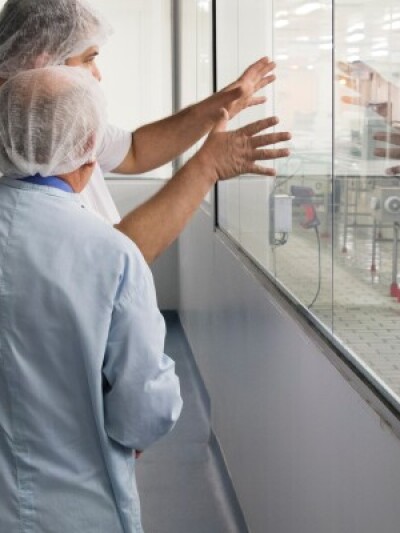 Two researchers in hairnets and lab coats looking through glass into a laboratory 