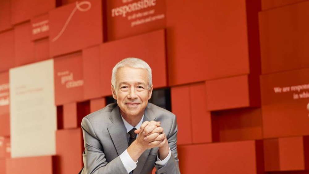 Johnson & Johnson CEO Joaquin Duato leaning on a table with his hands clasped in front of a red background 