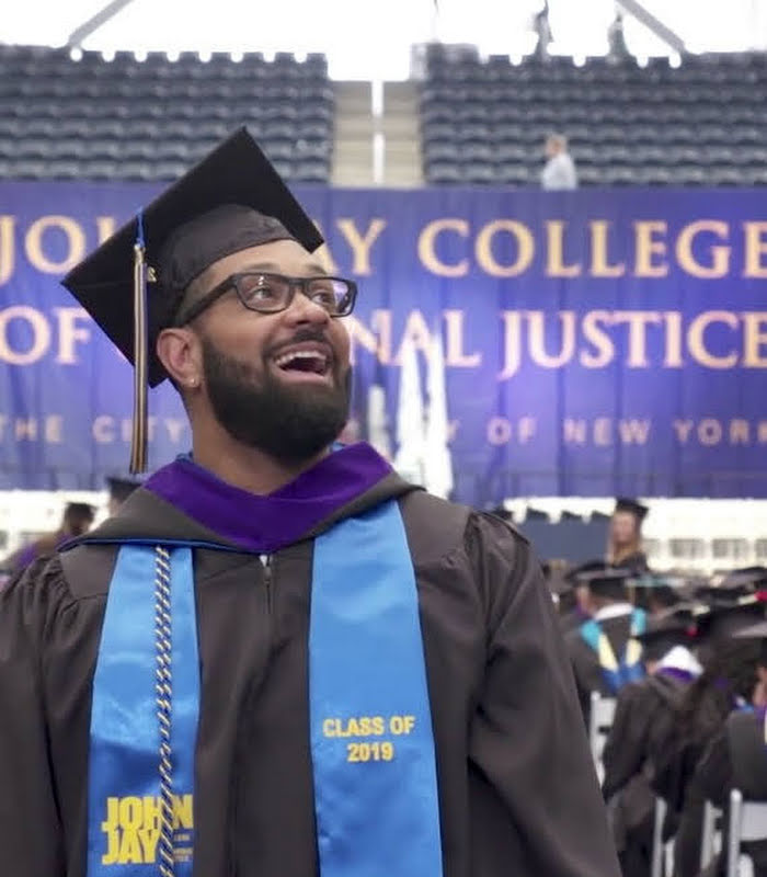 Excited John Jay College student dressed up in cap and gown, standing in auditorium at graduation ceremony