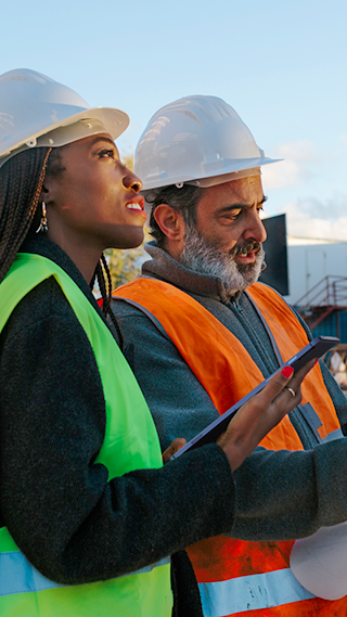 A Black woman wearing a hard hat and a yellow safety vest inspecting an outdoor job site using a mobile tablet with a Middle Eastern male colleague wearing a hard hat and an orange safety vest.