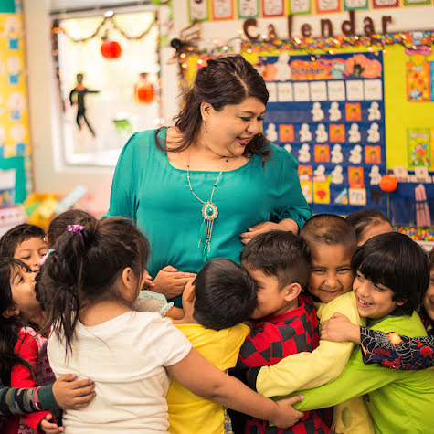 Smiling woman in a school classroom surrounded by a group of young children.