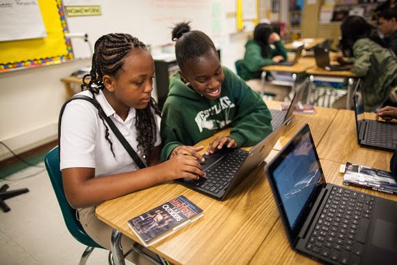 Dos niñas de secundaria comparten una computadora portátil en un salón de clases. Una de ellas, con un polo blanco, está concentrada en la pantalla. Su amiga, con una sudadera verde, sonríe a la pantalla.