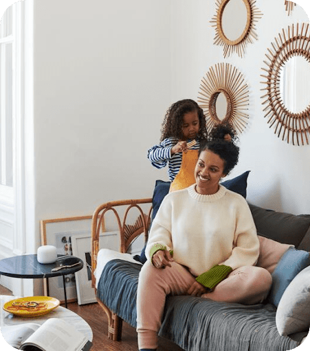 A little girl standing on a couch to help a smiling woman put her hair up with a barrette.