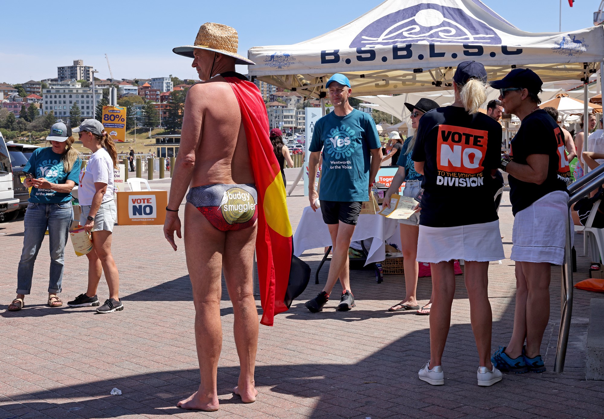 A man wearing Aboriginal flag speedos and a Aboriginal flag around his neck stands next No campaigners.