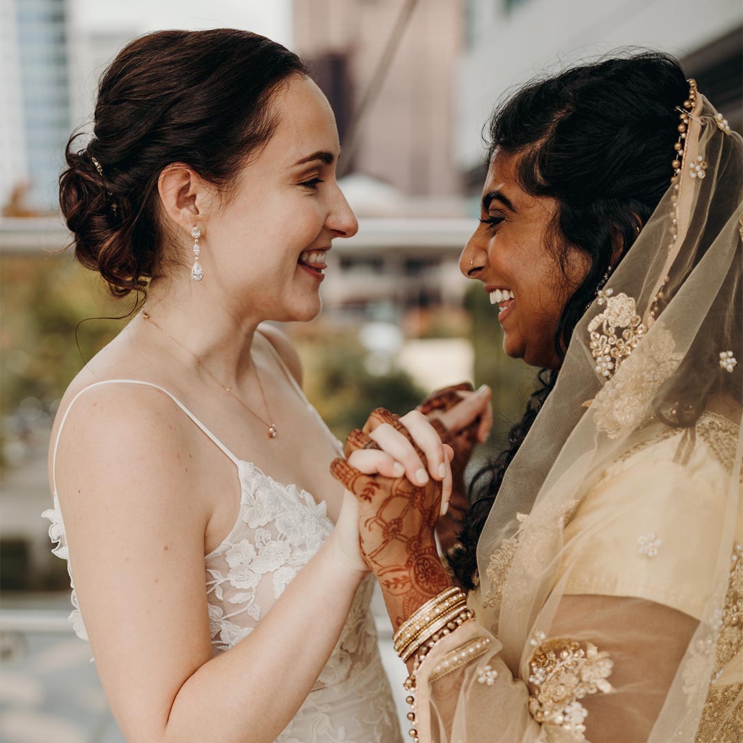 A wedding photo of two women outdoors, facing each other, smiling, and holding hands.