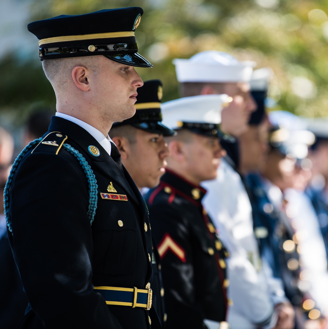 An Army soldier in dress uniform stands at attention with other service members, who are blurred into the background. A tree is also in the background.