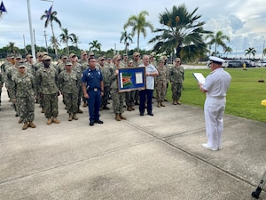 CNO Gilday reads a citation to a group of uniformed Sailors holding a plaque.