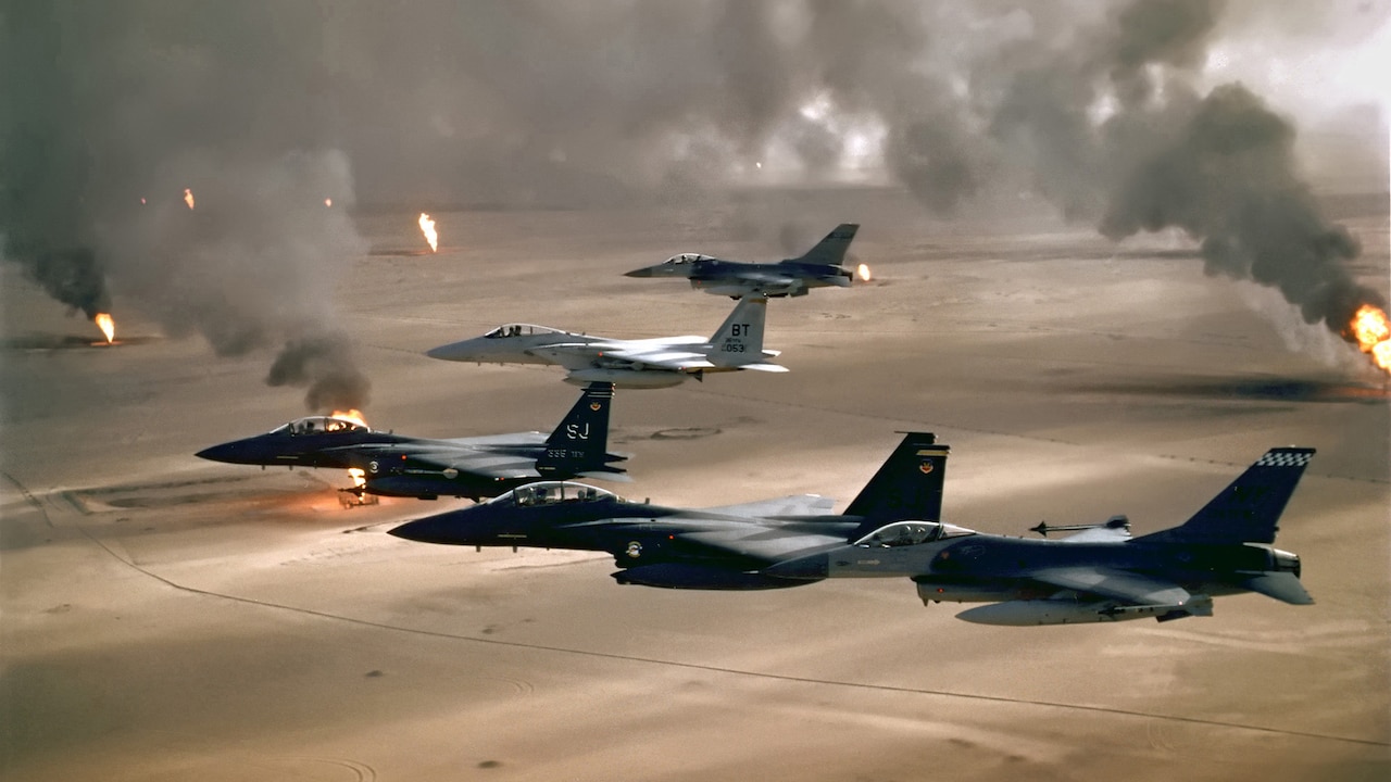 Jets fly in a V as detonations light the desert below.