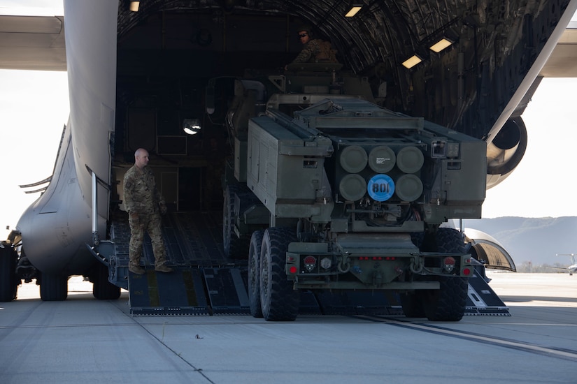 A military vehicle carrying several missiles rolls out of the back of an aircraft while a service member stands on the loading ramp.