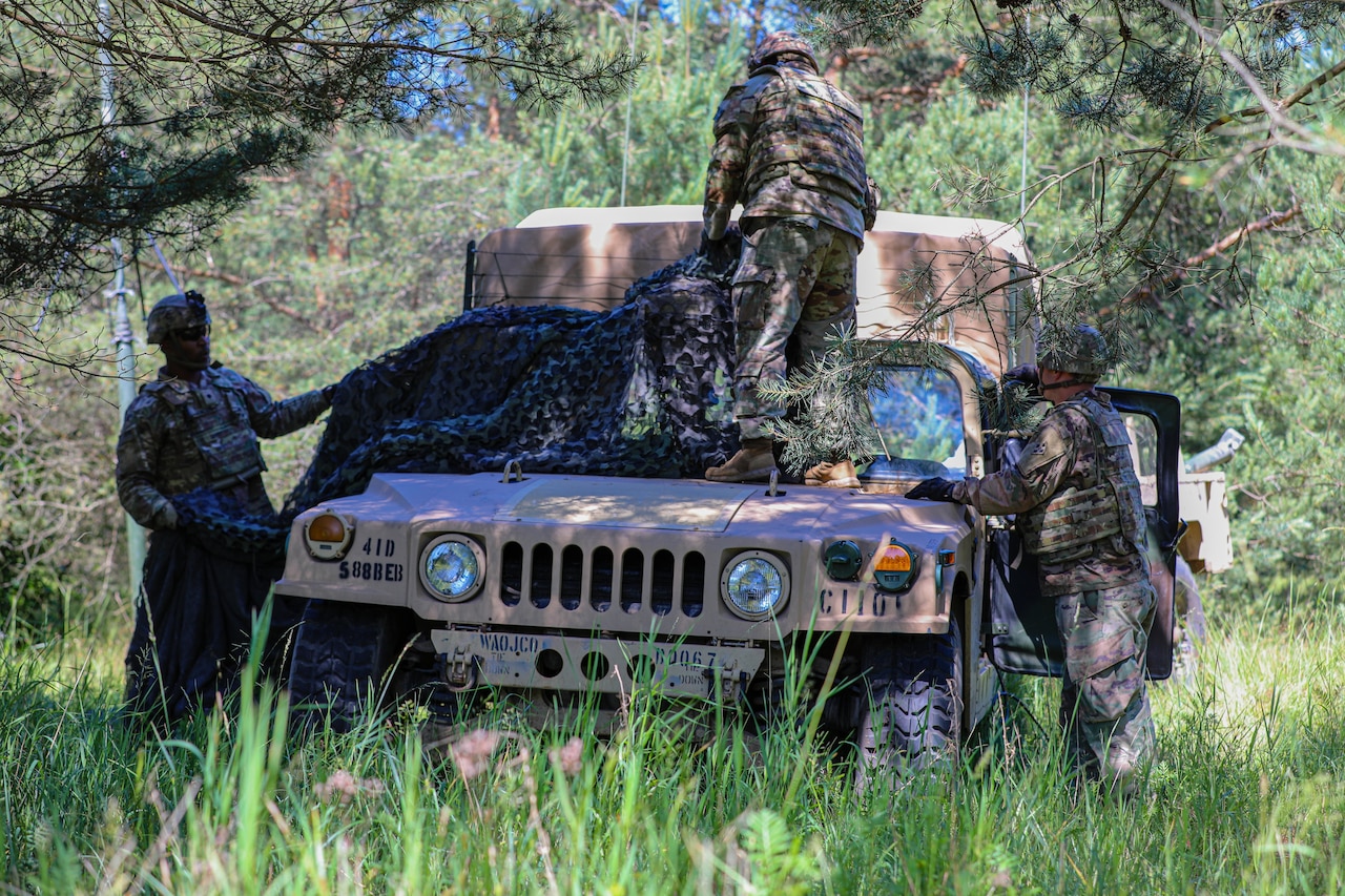 Three soldiers cover a Humvee with camouflage netting in a forested area.