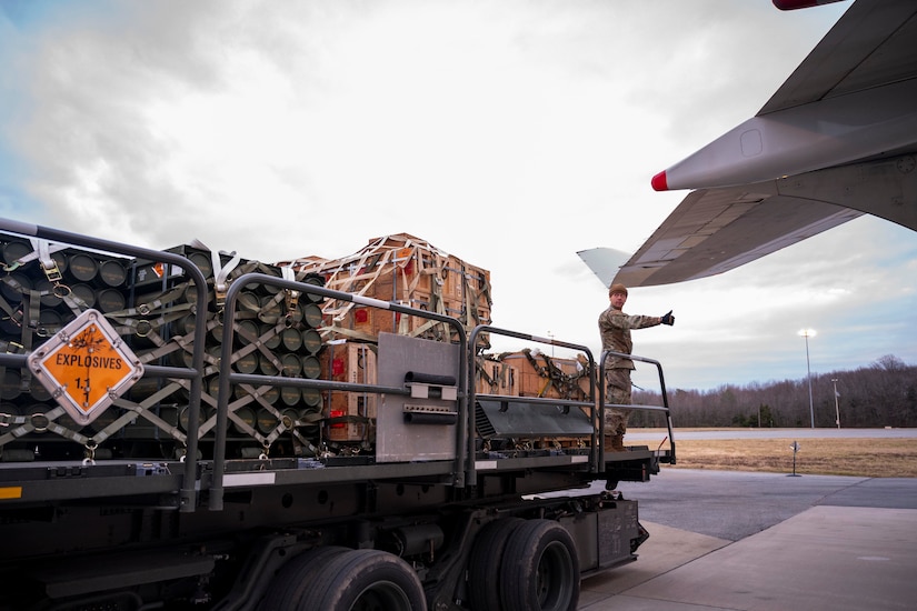 An airman in military uniform stands on a cargo loader parked near an airplane.