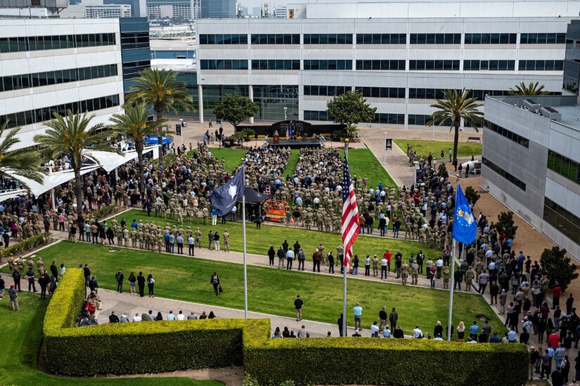 A group of people stand in a courtyard, facing a stage as a military leader addresses them.