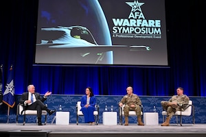 Four people, two in uniforms, sit in chairs on a stage. Behind them a projection screen has the words "AFA Warfare Symposium -- A Professional Development Event."