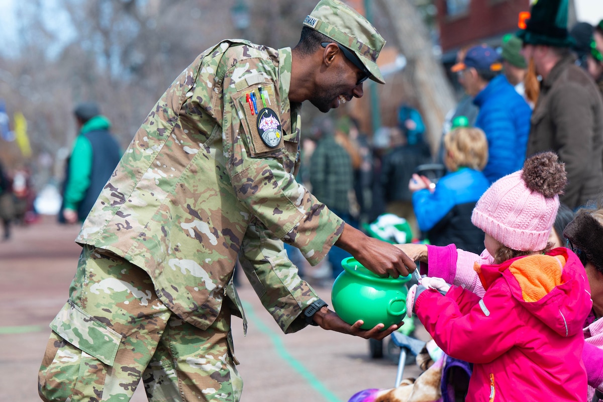 A guardian laughs as he holds out a green pot of candy for a child dressed in a hat and coat.