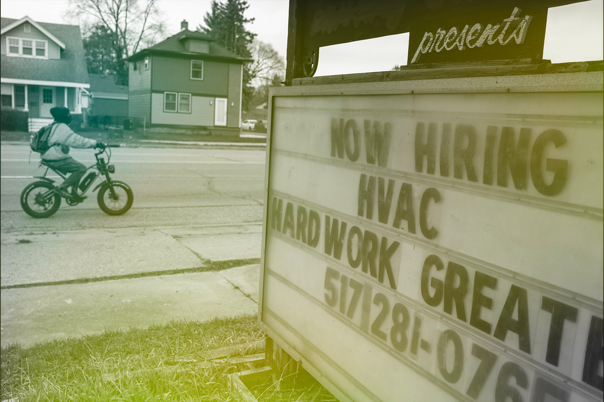 Person riding a bicycle past a sign that says NOW HIRING HVAC HARDWORK GREAT