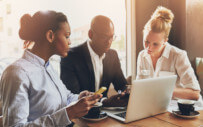 two women and a man working together at table on their phones and laptop