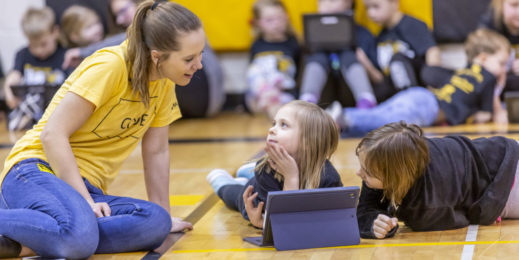 A young woman sits with two children as they use a tablet in a school gymnasium.