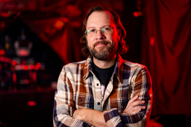 Mark Harnett stands with arms crossed in a dark lab lit with red lighting. 