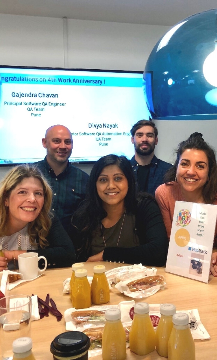 Five smiling PubMatic employees in an office with drinks and food around them. The text "Congratulation on 4th Work Anniversary!, Gajendra Chavan Principal Software AQ Engineer QA Team Pune, Divya Nayak Senior Software QA Automation Engineer QA Team Pune" behind them on a screen