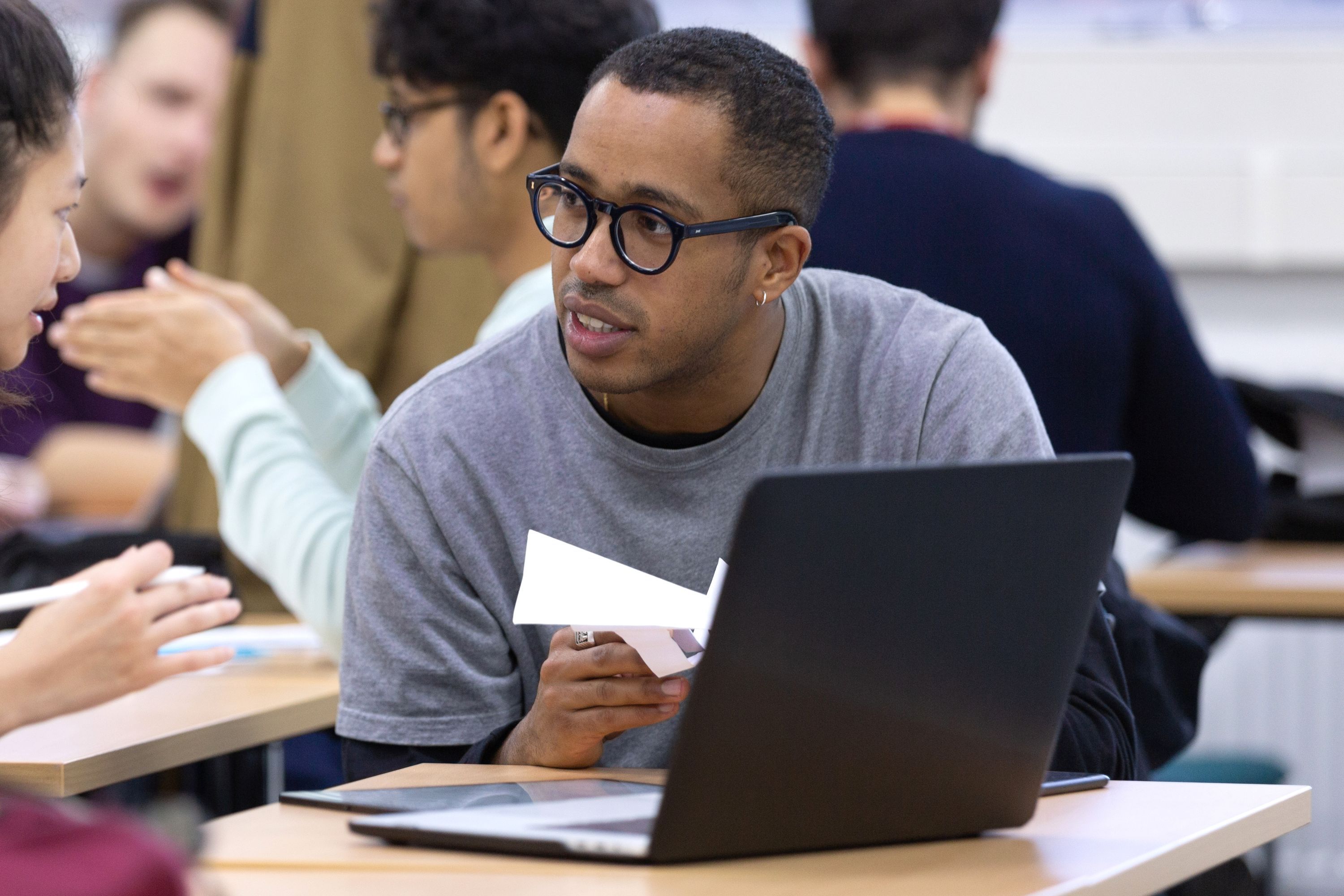 A male student sat at a laptop holding paper