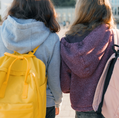 Two young girls from the back standing outside. One has a yellow backpack on, and the other has a light pink spotty backpack on.