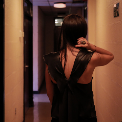 A young woman wearing a black dress stands in a hallway with her back to us.