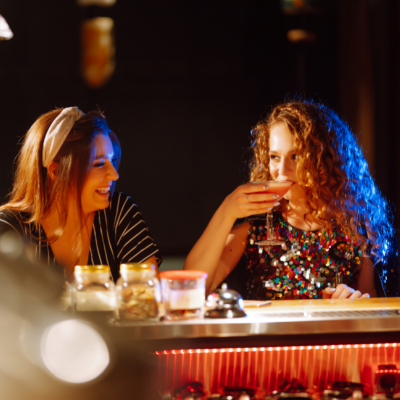 Two young women sit at a bar talking. One is sipping from a cocktail.