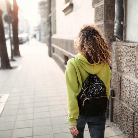 A young woman from the back, walking down the street with a backpack on in the sunshine.