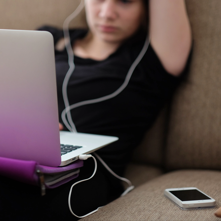 A young women with headphones in lies on a sofa, looking at her laptop.
