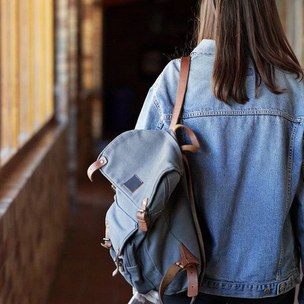 A teenage girl walks down a school corridor. She is wearing a denim jacket and has a backpack slung over one of her shoulders. The photo is taken from the back so we can't see her face.