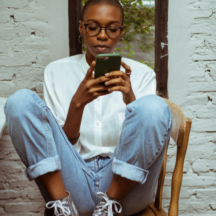 A young woman in round glasses sits on a kitchen chair using her smartphone. Her feet are up on the chair and she has her knees drawn up.