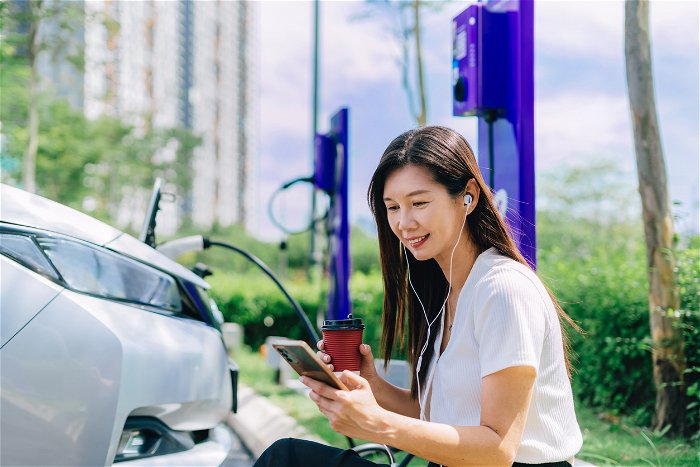 Asian woman chanring electric car outdoor, listening music and having coffee while waiting