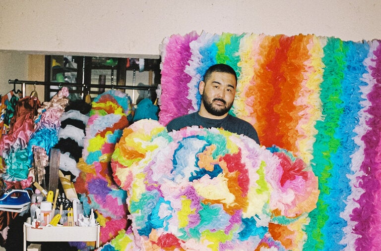 The fashion designer and artist Tomo Koizumi, photographed on July 11, 2023, at his dressmaking studio in Tokyo’s Shinjuku neighborhood with three of his art pieces made from leftover fabric: a giant teddy bear and, behind him, a pair of matching rainbow banners.