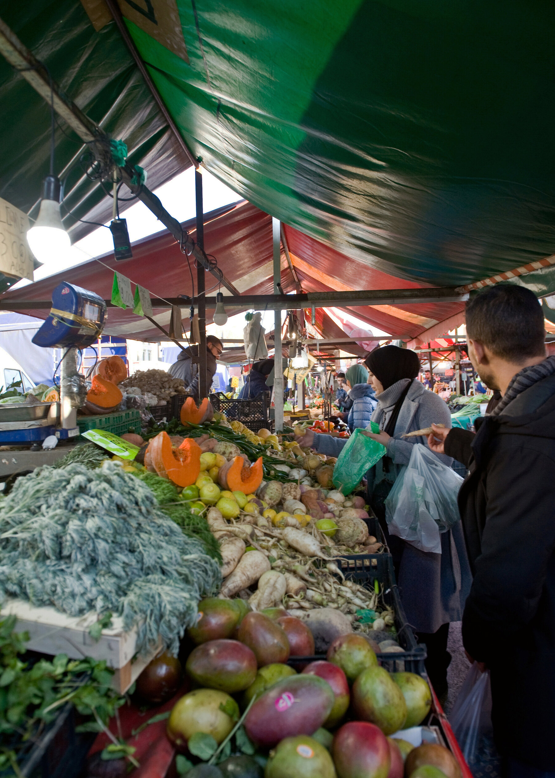 People shop for fresh produce (including mangoes, pumpkin and root vegetables) in an open-air market with a tarp canopy. 